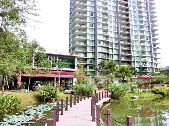 A wooden walkway leads through a pond with lily pads towards a modern high-rise apartment building and a garden cafe.