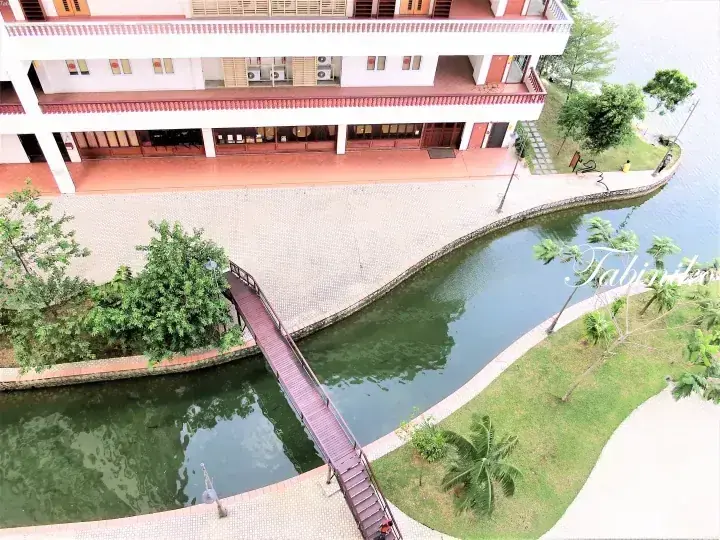 Aerial view of a resort building beside a canal with a wooden bridge, palm trees, and a grassy bank.