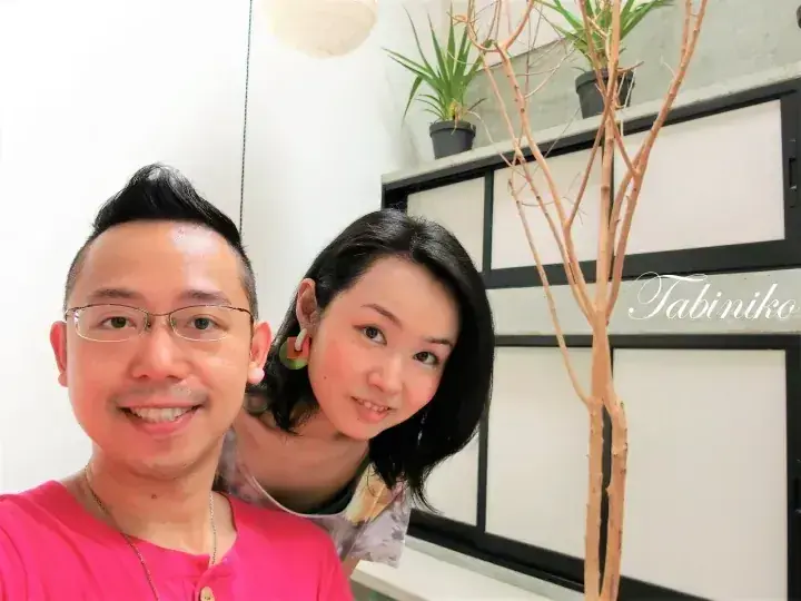A couple poses for a selfie in a modern interior with plants, featuring the Tabiniko watermark.