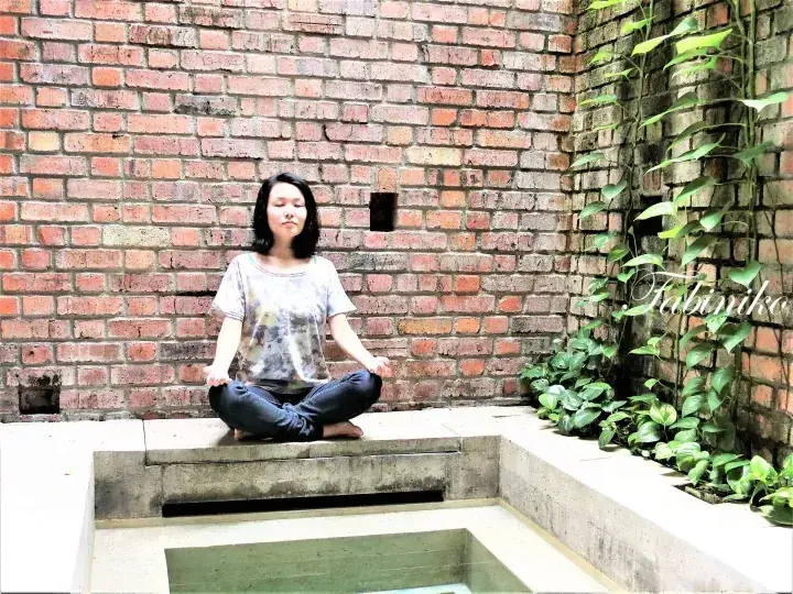 A woman meditating in a lotus pose at a serene courtyard with exposed brick walls and climbing greenery.