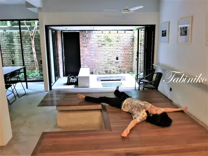A woman lies on the wooden floor of a modern home with open sliding doors revealing a brick courtyard.
