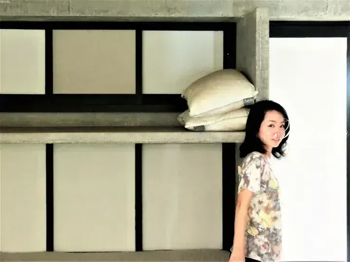 Woman standing beside a concrete shelf unit with rice sacks in a minimalist interior.