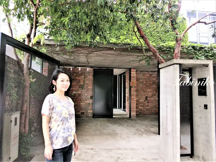 赤レンガとコンクリートのモダンな住宅の入り口で緑に囲まれた女性。