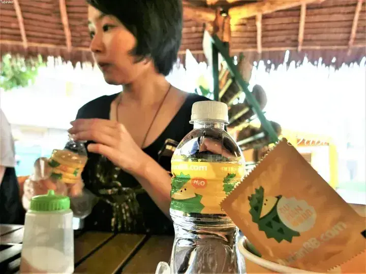 Woman at a table with DurianBB.com branded water bottle and snack bag in a rustic open-air setting.