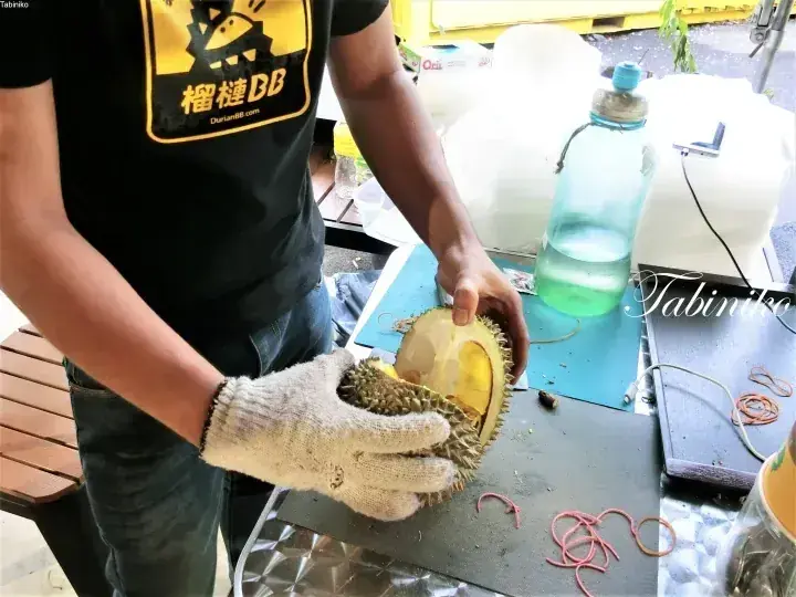 DurianBB vendor opens a durian fruit at a stall, wearing protective gloves and a branded shirt.