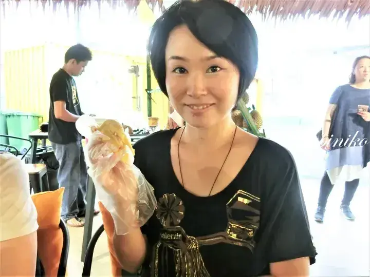 Smiling woman holding a plastic bag of durian at an open-air stall.