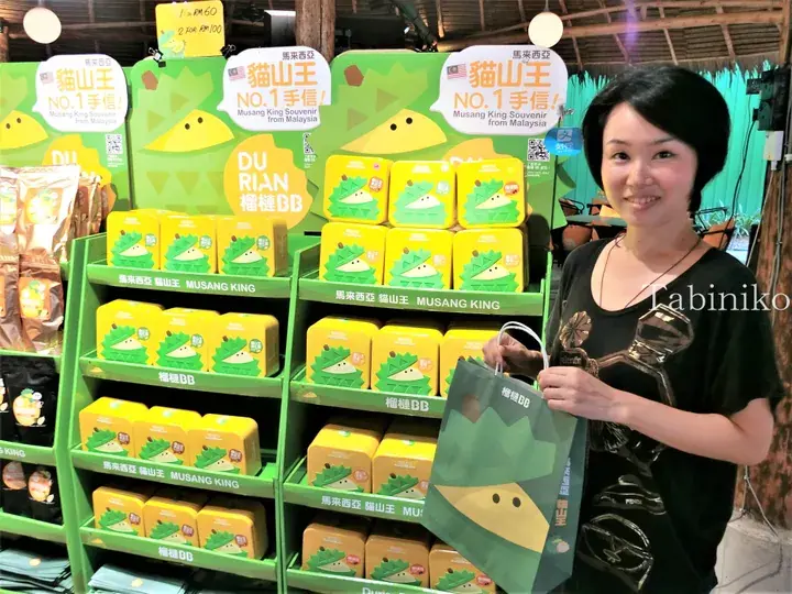 マレーシアの店で、猫山王のドリアン土産が並ぶディスプレイの前にDurianBBのバッグを持った女性が立っている。