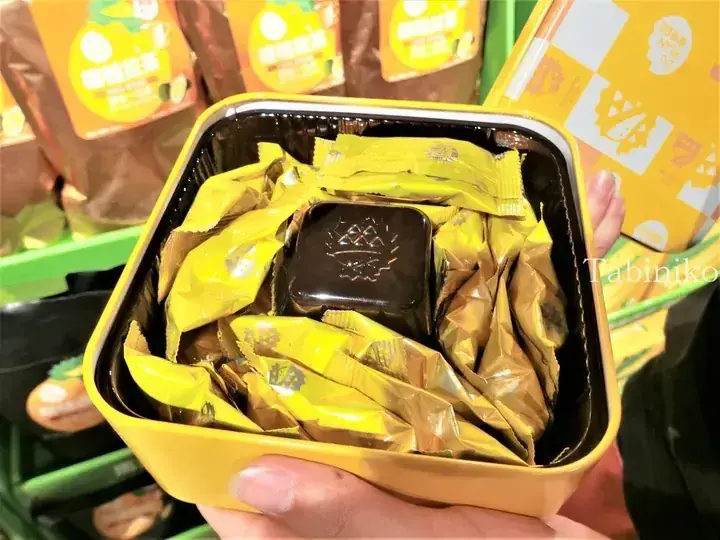 Yellow tin filled with individually wrapped chocolates and a central durian-shaped chocolate, with Durian Latte packaging in the background.