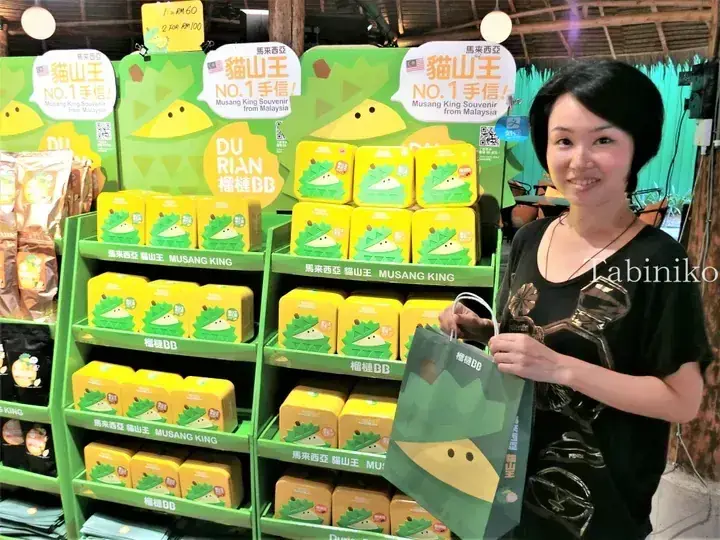 A woman holding a DurianBB bag stands in front of a display of Musang King durian souvenirs at a Malaysian shop.