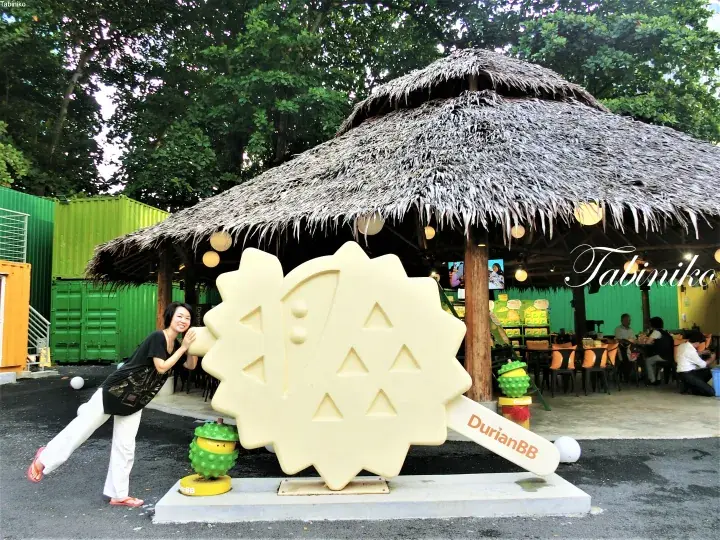 茅葺き屋根の屋外飲食店「DurianBB」で、巨大なドリアンアイスクリームのオブジェと記念撮影する女性。