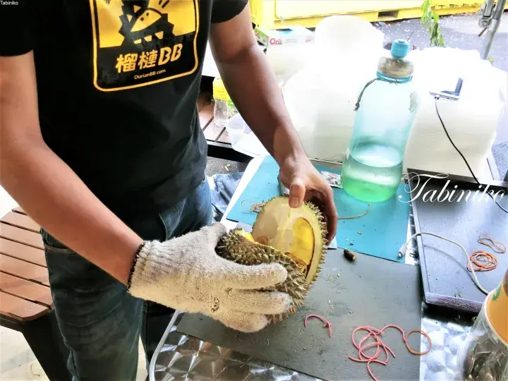 防護手袋とブランドTシャツを着た店員が、屋台でドリアンBBのドリアンを割っている様子。