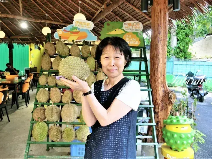 Woman holding a durian in front of a display rack at a Malaysian durian stall.