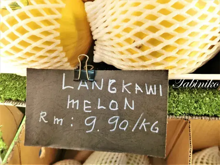 Price sign for Langkawi Melon at RM 9.90/kg displayed in front of yellow melons wrapped in protective foam netting.