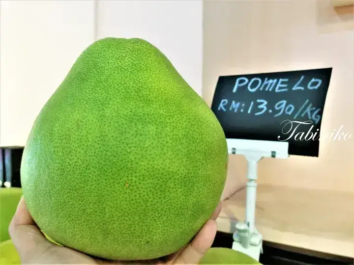 Hand holding a large green pomelo with a price sign showing RM13.90/kg in the background.