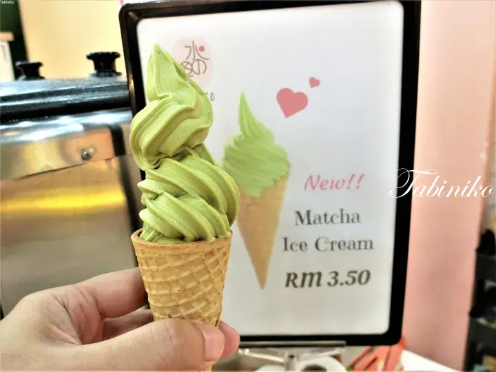 背景に「Matcha Ice Cream RM 3.50」と書かれた看板を映しながら、抹茶ソフトクリームを手に持っている様子。