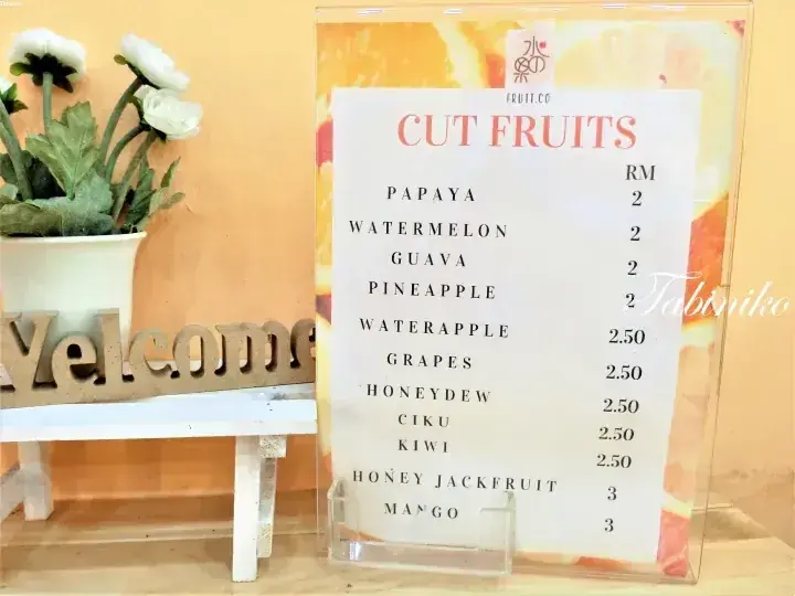 Fruit.co cut fruits menu with prices in RM, displayed next to a white pot and wooden welcome sign.