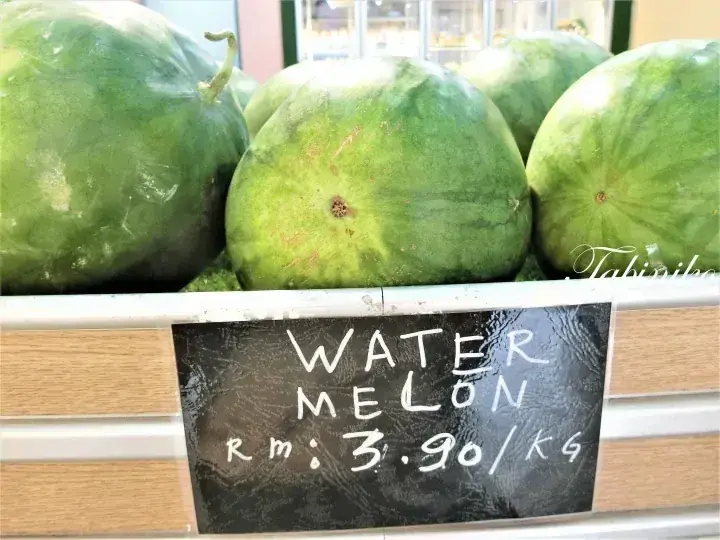 Fresh watermelons displayed on a supermarket shelf with a chalkboard sign showing the price RM 3.90 per kg.
