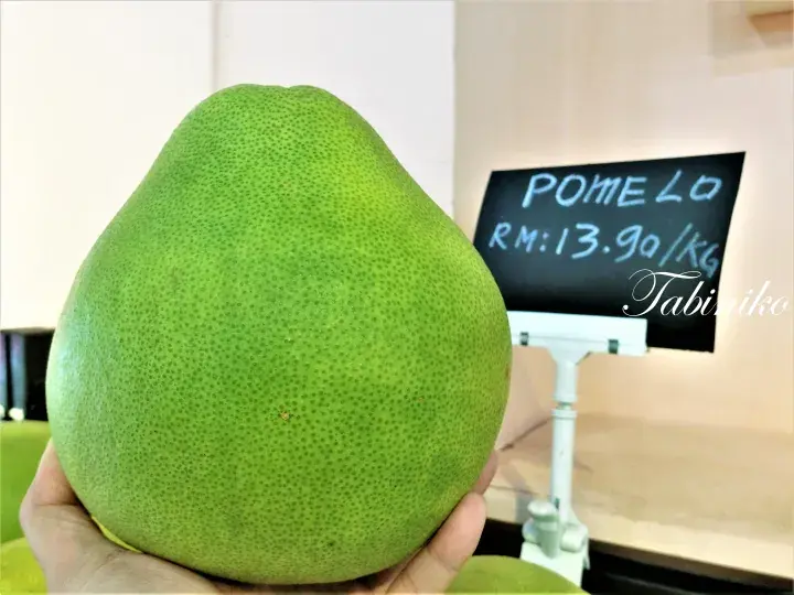 背景に「RM13.90/kg」の価格表示がある緑色の大きなポメロを手に持つ様子。