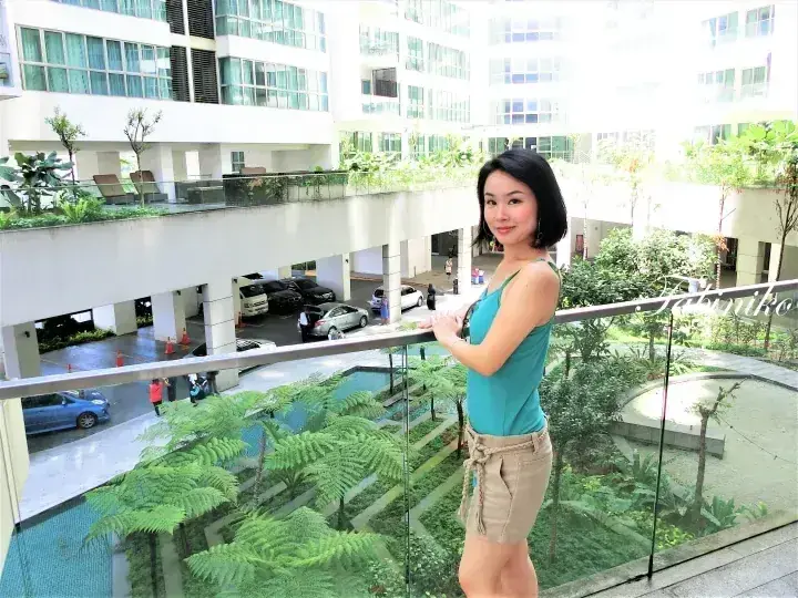 Woman in teal top and beige shorts stands on a balcony overlooking a lush courtyard with ferns and a modern hotel building.