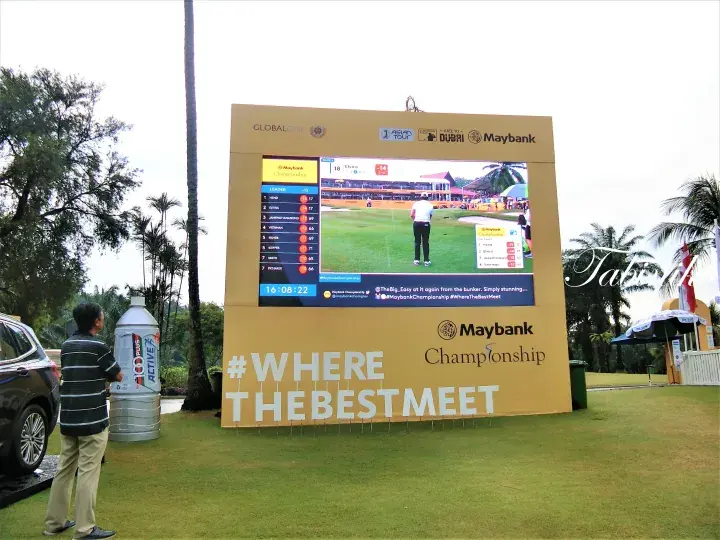 A large Maybank Championship scoreboard displays live golf action and scores at a lush green course.