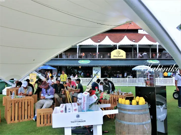 Guests relax under a white tent at a Maybank golf event, with Taittinger branding and wooden crates visible.