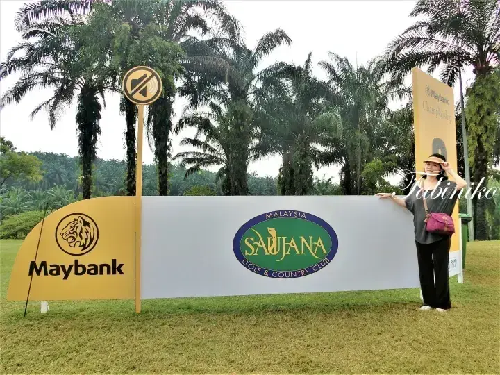 パームツリーを背景に、Maybank Saujana Golf & Country Clubの看板の前でポーズをとる女性。