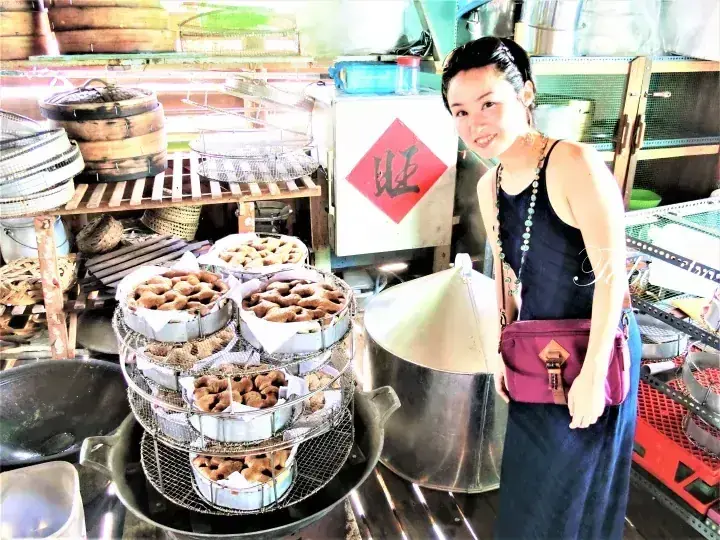 A woman stands beside a large steamer filled with traditional Malaysian kuih at a local food stall.