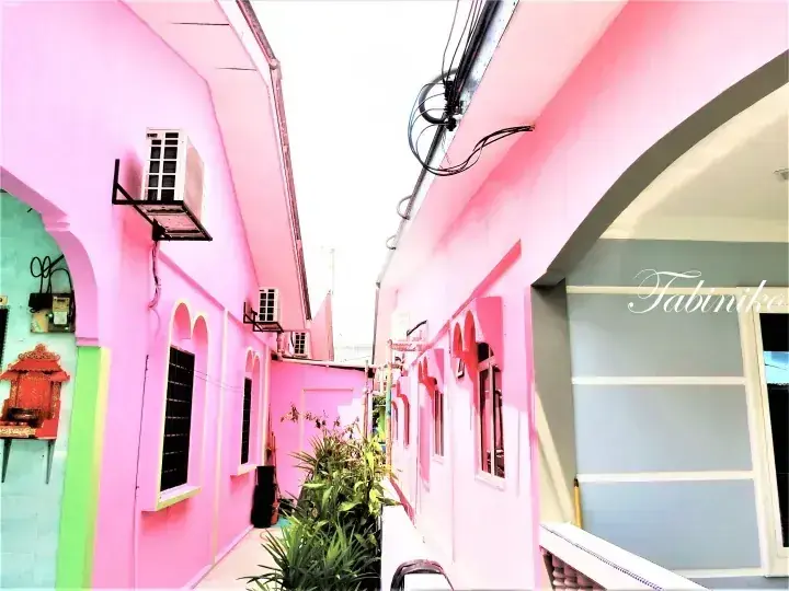 Narrow alleyway between pink buildings at Tabinka, featuring arched windows, air conditioning units, and a small spirit house.