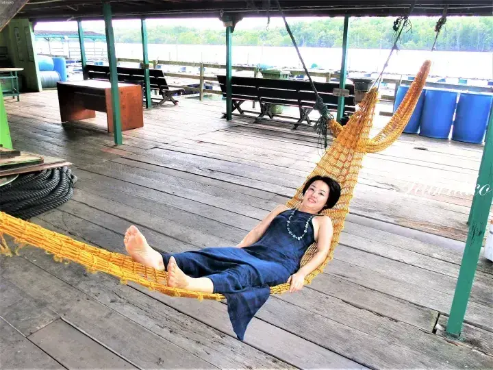 Woman relaxing in a woven hammock on a wooden deck overlooking a river in Malaysia.