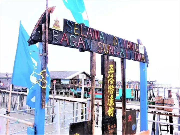 Welcome sign reading SELAMAT DATANG BAGAN SUNGAI LIMA at a traditional stilt village with blue flags and wooden structures.