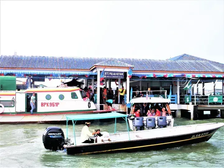 Boats docked at Jeti Pulau Ketam jetty with passengers boarding and a speedboat passing by in the foreground.