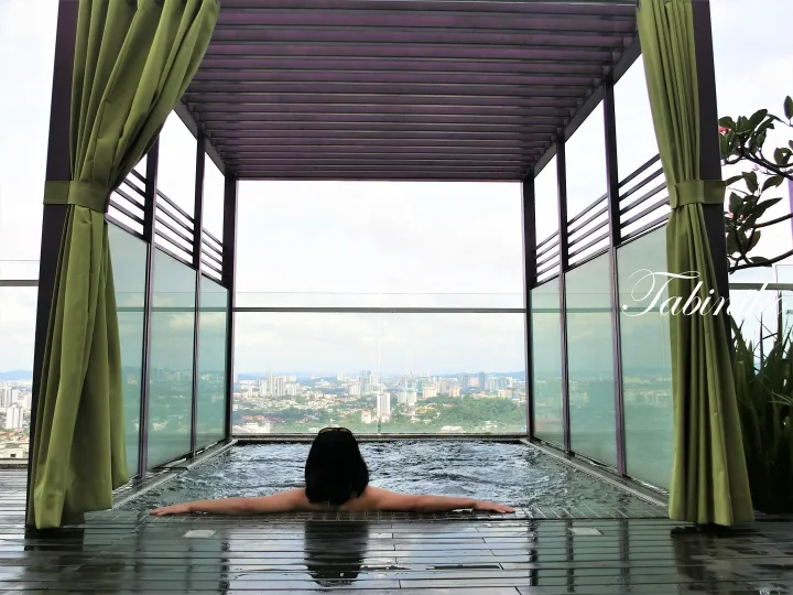Person relaxing in a rooftop infinity pool with a city view, framed by green curtains and a wooden pergola.