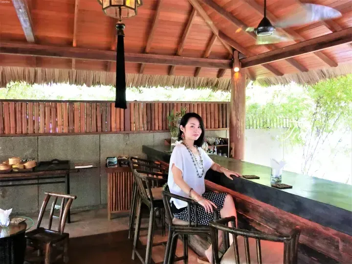 Woman sitting at a wooden bar counter inside a rustic open-air pavilion with bamboo walls and a thatched roof.