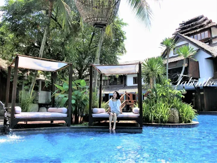 Woman relaxing on a daybed by a pool at a tropical resort with thatched roofs and lush greenery.
