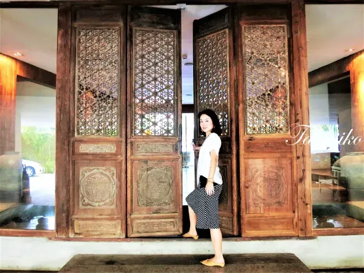 A woman stands in the ornate wooden doorway of a heritage property, showcasing intricate lattice carvings and traditional design elements.