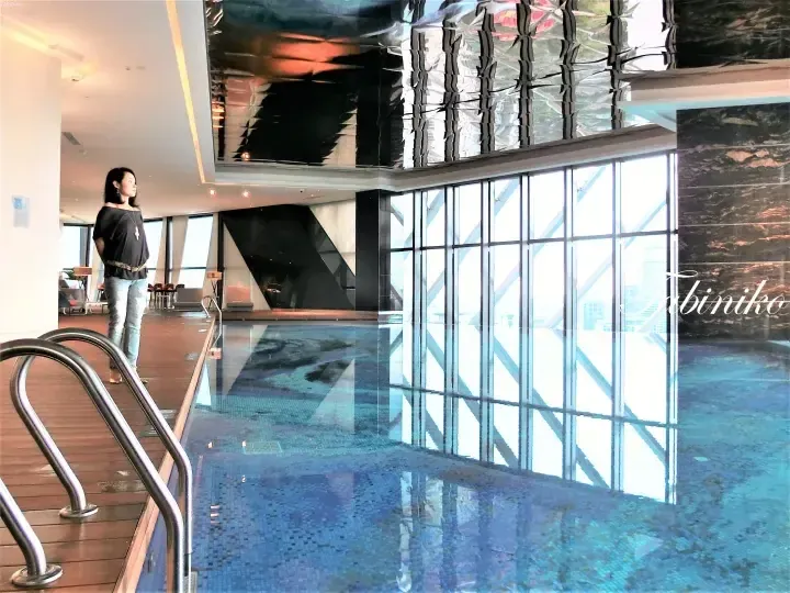 Woman standing by indoor infinity pool with city views at The Pearl Kuala Lumpur.