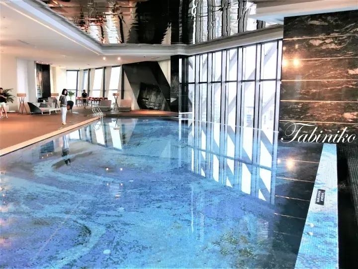 Infinity pool with blue mosaic tiles at a high-rise hotel, featuring floor-to-ceiling windows and a dark marble wall.