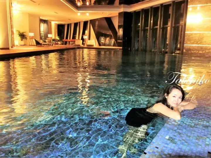 Woman relaxing in an indoor pool at night with warm lighting and lounge seating visible in the background.