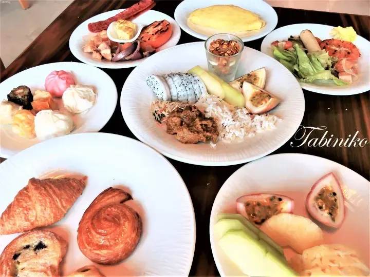 Assorted breakfast buffet spread including pastries, dim sum, omelette, grilled vegetables, and tropical fruits on white plates.