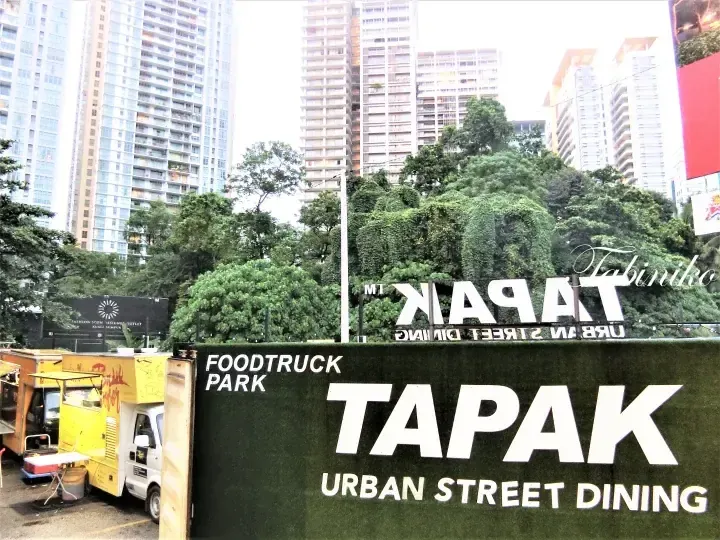 TAPAK Urban Street Dining food truck park entrance with yellow food trucks and high-rise buildings in the background.