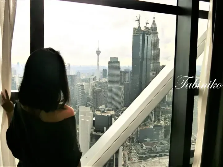 Woman looking out a high-rise window at the Kuala Lumpur skyline featuring the Petronas Twin Towers and KL Tower.