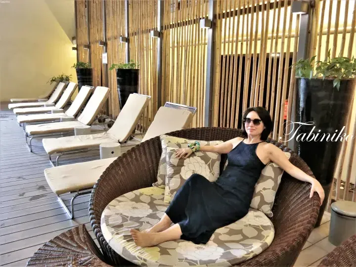 Woman relaxing in a round wicker chair at a resort lounge with bamboo screens and sun loungers.