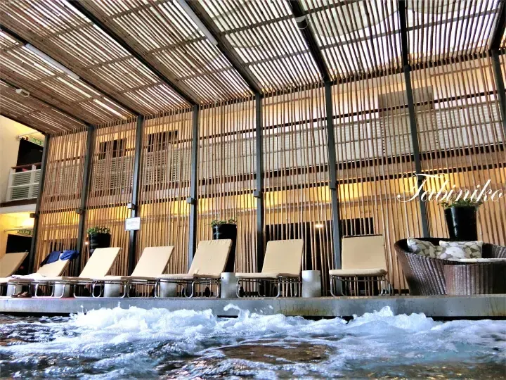 Lounge chairs beside a bubbling pool under a bamboo slat roof at a resort.