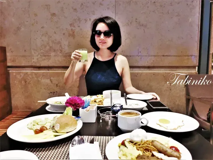Woman in sunglasses and black swimsuit holding a green drink at a breakfast buffet table with various dishes.