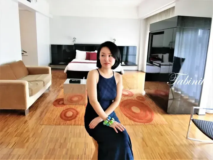 Woman in a blue dress sitting on a stool in a modern hotel suite with a bed, sofa, and wooden flooring.
