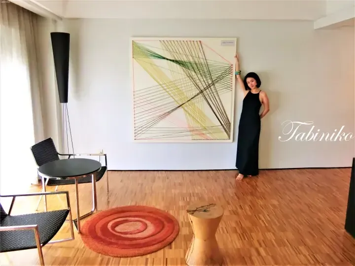 Woman in black dress posing beside a large string art piece in a modern hotel suite with wooden floors and contemporary furniture.