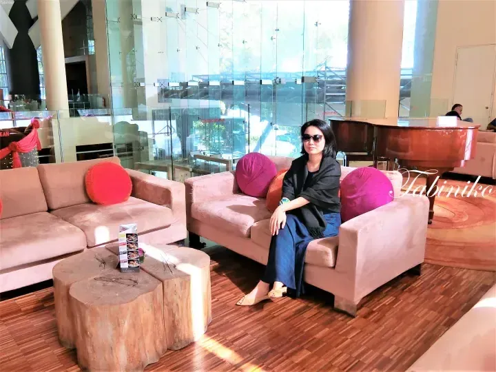 Woman in sunglasses sitting on a pink sofa in a modern hotel lobby with a grand piano and wooden coffee tables.