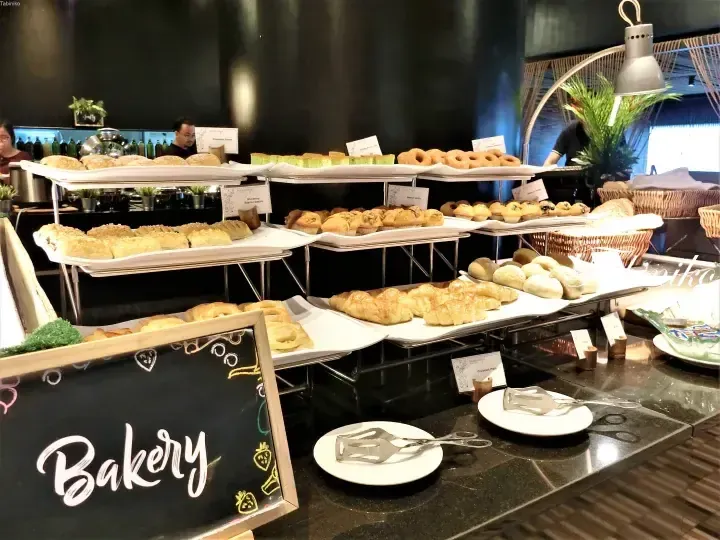Hotel Maya buffet bakery station displaying croissants, muffins, danish pastries, and breads on tiered racks with a chalkboard sign.