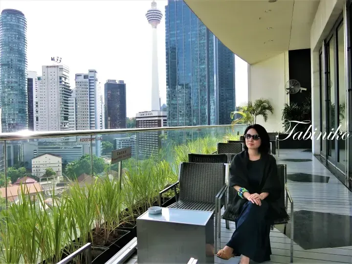 Woman relaxing on a balcony with a view of Kuala Lumpur skyline, featuring the KL Tower and KL33 building.