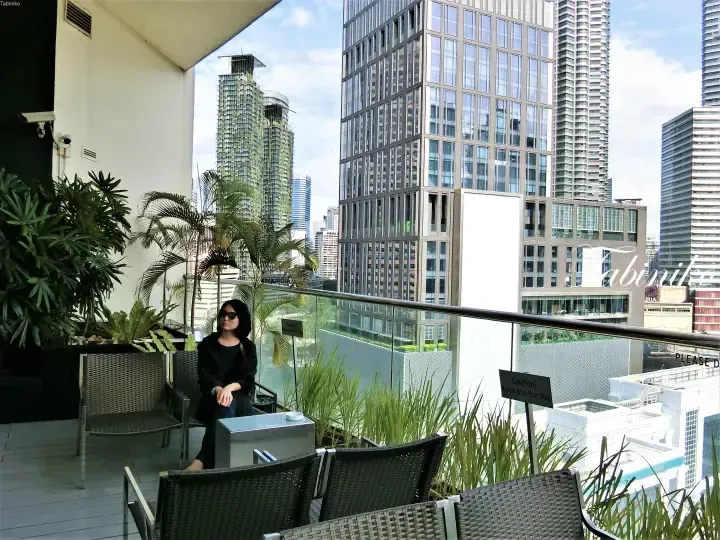 Woman relaxing on a balcony terrace overlooking the Kuala Lumpur city skyline with modern skyscrapers.